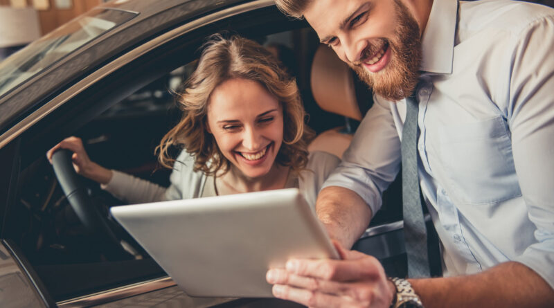 Schöne junge Frau unterhält sich mit dem gutaussehenden bärtigen Verkaufsleiter bei der Auswahl eines Autos im Autohaus. Der Mann benutzt ein digitales Tablet