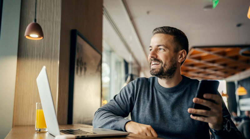 Ein glücklicher Freiberufler arbeitet in einem Café an sozialen Medien und nutzt dabei mehrere Technologien.